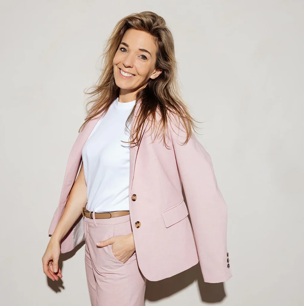 Een persoon in een roze blazer die glimlachend poseert tegen een neutrale achtergrond.