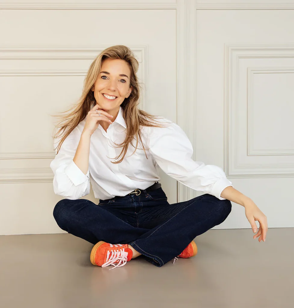 Persoon met lang blond haar, zittend op de vloer in een witte blouse, jeans en rode schoenen, glimlachend naar de camera.