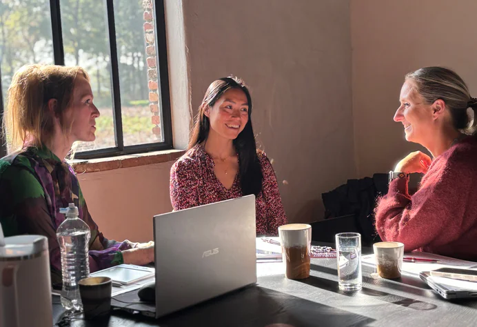 Twee vrouwen overleggen aan een tafel met een laptop en drinken bij daglicht.